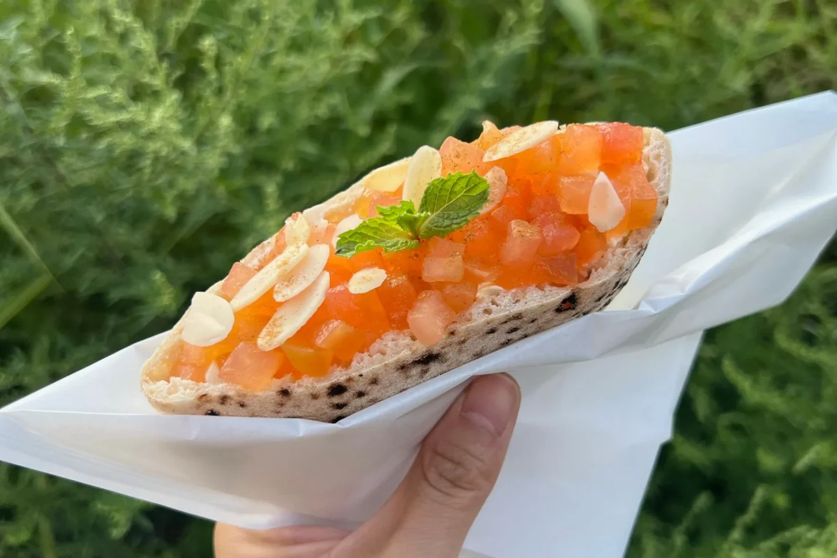 トマトとミントが乗ったおしゃれなオープンサンドが、白い紙に包まれて手で持たれている写真です。背景には緑色のぼやけた草木があります。