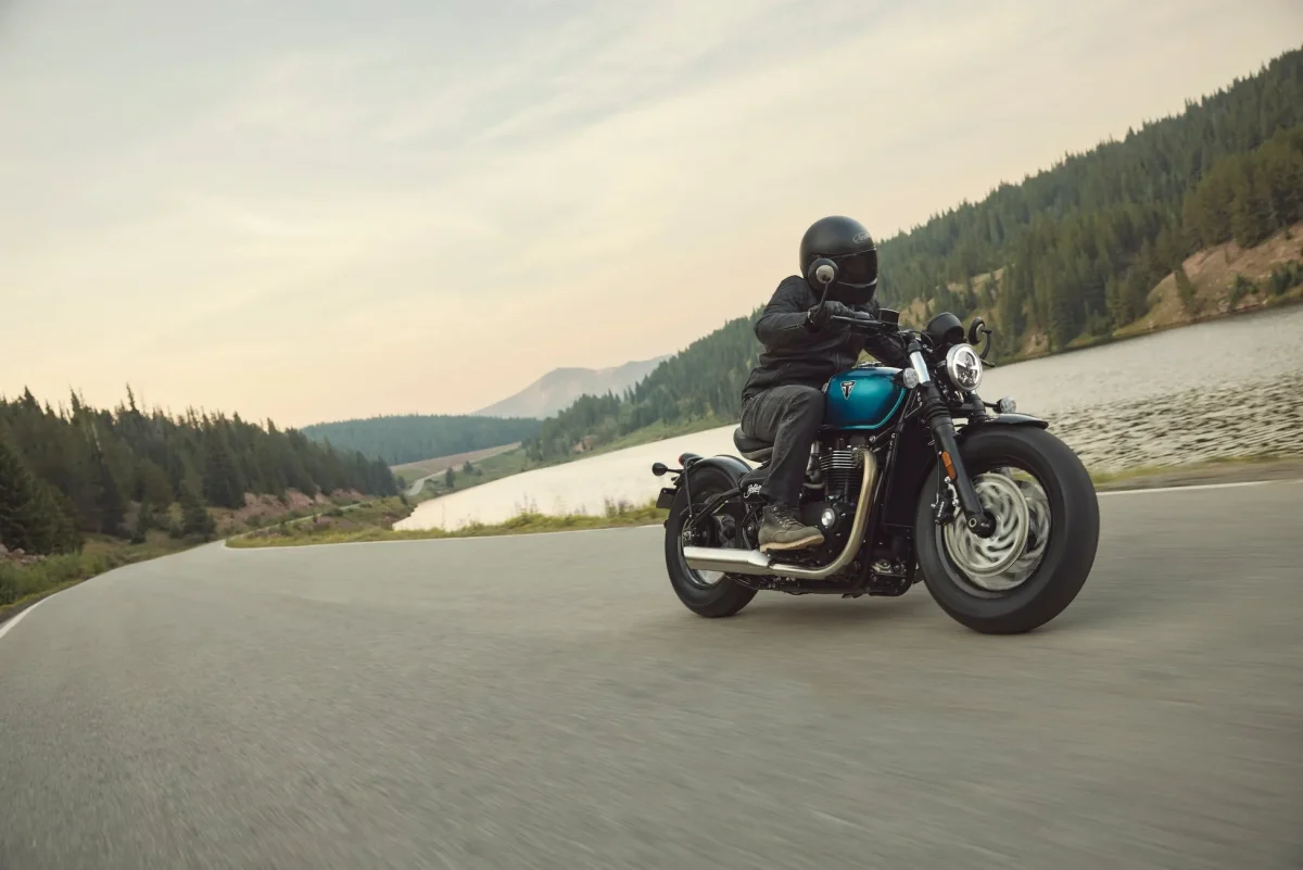 Triumphのバイクに乗って道路を走る人、背景には山と水面