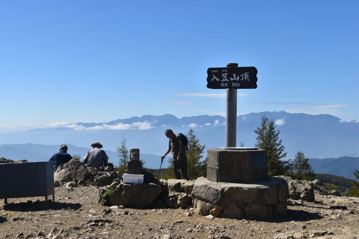 入笠山の山頂付近で、標識や風景を眺める人々の様子を捉えた写真です。青空の下、山頂を示す標識が設置され、登山客が休憩しています。遠くには山々が連なり、自然豊かな風景が広がっています。