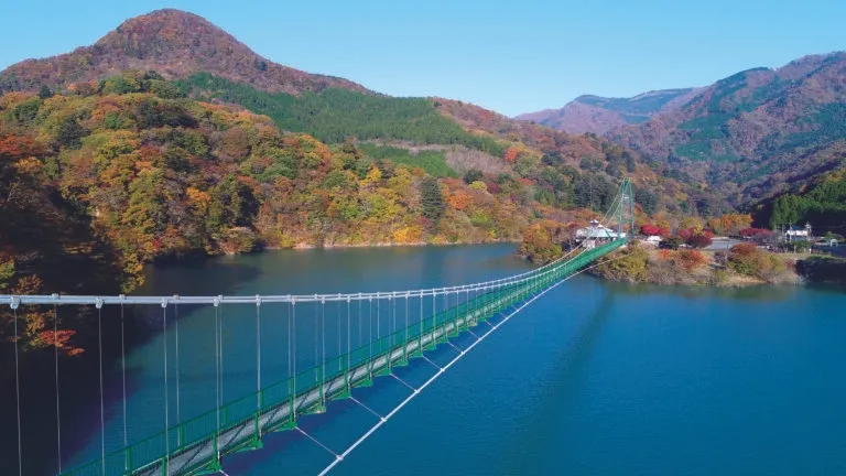 湖にかかる吊り橋の風景。周囲には紅葉した山々が広がり、美しい景色を作り出している。